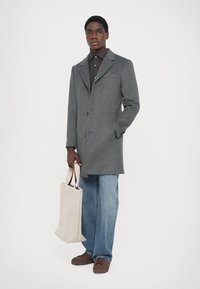 Grå ullkappa med slagkrage, en knapp och framfickor. Matchad med en brun skjorta, ljusblå jeans och en beige weekendväska.