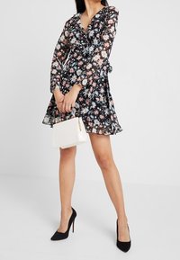 Vestido negro con flores, de manga larga y cuello con volantes. Presenta coloridos patrones de flores. Un bolso blanco y unos tacones altos negros completan el look.