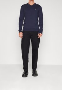 Pull col V bleu marine, tissu tricot doux ; associé à un pantalon noir ample et des chaussures noires à enfiler ; design minimaliste, sans motifs visibles.