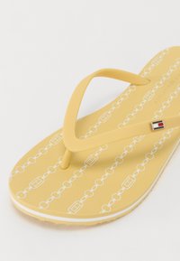 Tongs jaunes avec motif en chaîne blanche, dotées d'une semelle texturée et de brides souples et flexibles. Accentées d'un petit détail de logo.