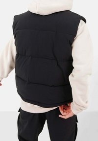 Gepolsterte schwarze Weste mit hohem Kragen, die horizontale Nähte und eine glatte Stoffstruktur aufweist, über einem hellen Hoodie getragen.