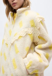 Κοντινό πλάνο ενός ατόμου που φοράει ένα κρεμ φλις μπουφάν με κίτρινα διαμάντια και δαντελένια διακόσμηση κατά μήκος του στήθους και των ώμων.