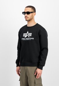 Uomo che indossa una felpa nera Alpha Industries con logo e pantaloni cargo oliva, in piedi contro uno sfondo bianco semplice.