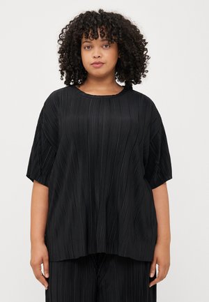 A person with curly hair wears a black pleated top, standing against a light background, looking directly at the viewer.