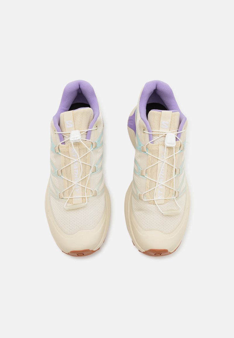 Par de zapatillas deportivas beige con forro interior morado, acentos laterales azul claros y sistema de cordones rápidos blancos sobre un fondo blanco.