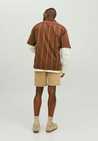 Braunes Kurzarmhemd mit vertikalen Streifen, kombiniert mit beigen Shorts. Das Hemd hat einen Kragen, und das Outfit umfasst hellfarbene Schuhe und Socken.
