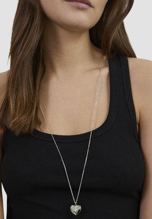 Silberne Herz-Halskette mit einem Diamantakzent, an einer feinen Kette hängend, getragen über einem schwarzen, gerippten Tanktop.