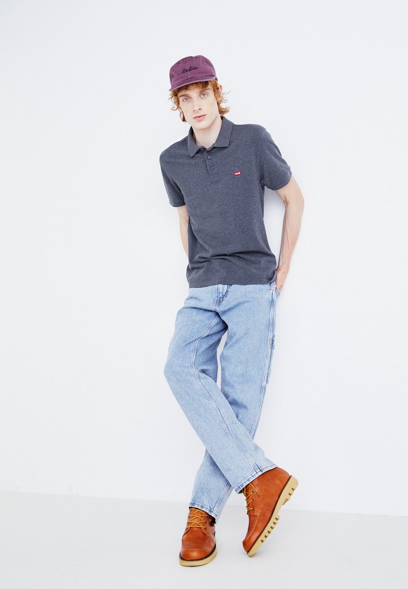 Polo de color gris oscuro con un pequeño logo rojo, jeans de color azul claro, botas de cuero marrón y una gorra morada, presentados sobre un fondo blanco.