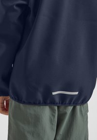 Navy windbreaker with smooth texture, featuring a white horizontal accent at the hem. Paired with light green, loose-fit pants.