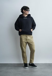 Jauns vīrietis melnā džemperī ar kapuci, bēšās pieguļošās biksēs, melnās sporta kurpēs un saulesbrillēs stāv pie vienkāršas gaiši pelēkas sienas.