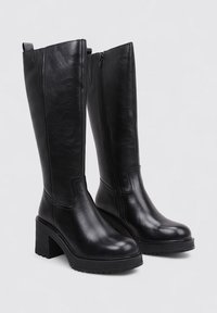 Bottes en cuir noir jusqu'au genou avec un talon épais et une semelle texturée, dotées d'une fermeture éclair sur le côté pour un enfilage facile et de détails de couture minimaux.