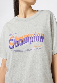 Camiseta de algodón gris con mangas cortas que presenta el logo de "Champion" en texto púrpura y naranja en el pecho. Cuello redondo, corte casual.