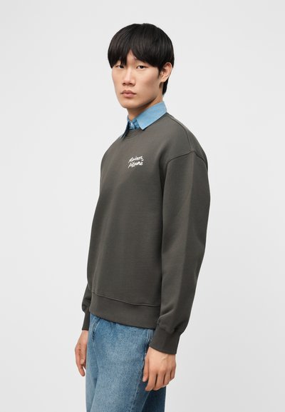 Νέος άνδρας που φοράει σκούρο γκρι φούτερ πάνω από μπλε πουκάμισο με γιακά και ανοιχτόχρωμο τζιν, στέκεται μπροστά σε απλό φωτεινό φόντο.