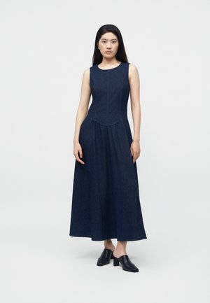 Femme aux longs cheveux foncés debout face à l'objectif, portant une robe maxi bleu marine sans manches et des mules à talons noires, sur un fond uni clair.