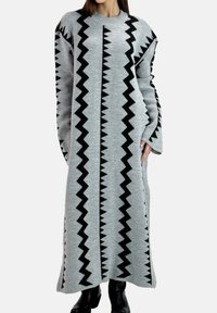 Grå strikket kjole med et zigzag-sort mønster langs siderne, lange ærmer og en løs silhuet. Tekstureret stof med rund halsudskæring.