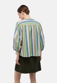 Blouse rayée en tissu léger aux teintes bleu, vert, jaune et orange. Manches bouffantes et coupe décontractée, portée sur une jupe vert foncé.