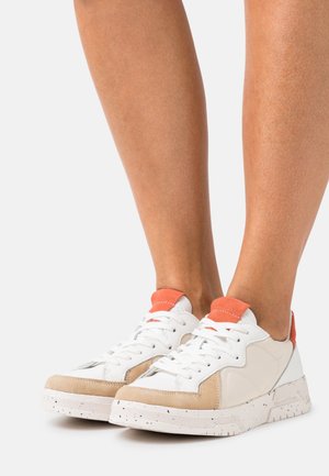 Zapatillas deportivas de encaje blancas y beige con detalles naranjas en el talón y la lengüeta, usadas sobre piernas desnudas contra un fondo liso.