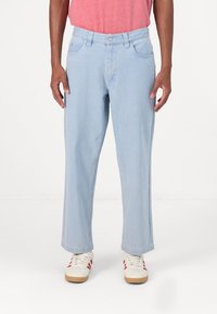 Ανοιχτό μπλε τζιν παντελόνι με ίσια γραμμή, πέντε τσέπες και διακριτική ραφή. Φοριέται με λευκά αθλητικά παπούτσια με κόκκινες λεπτομέρειες.