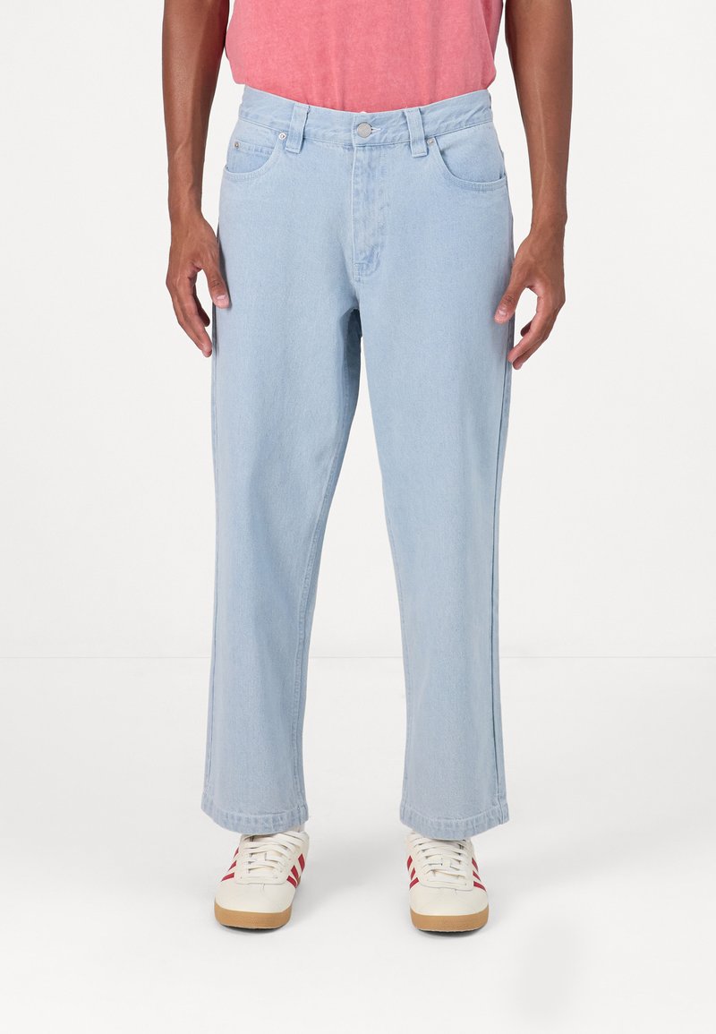 Ανοιχτό μπλε τζιν παντελόνι με ίσια γραμμή, πέντε τσέπες και διακριτική ραφή. Φοριέται με λευκά αθλητικά παπούτσια με κόκκινες λεπτομέρειες.