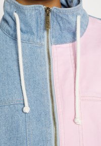 Džínová bunda s tělem v světle modré barvě a růžovým panelem, vysokým límcem, bílými stahovacími šňůrkami a viditelným zlatým zipem.
