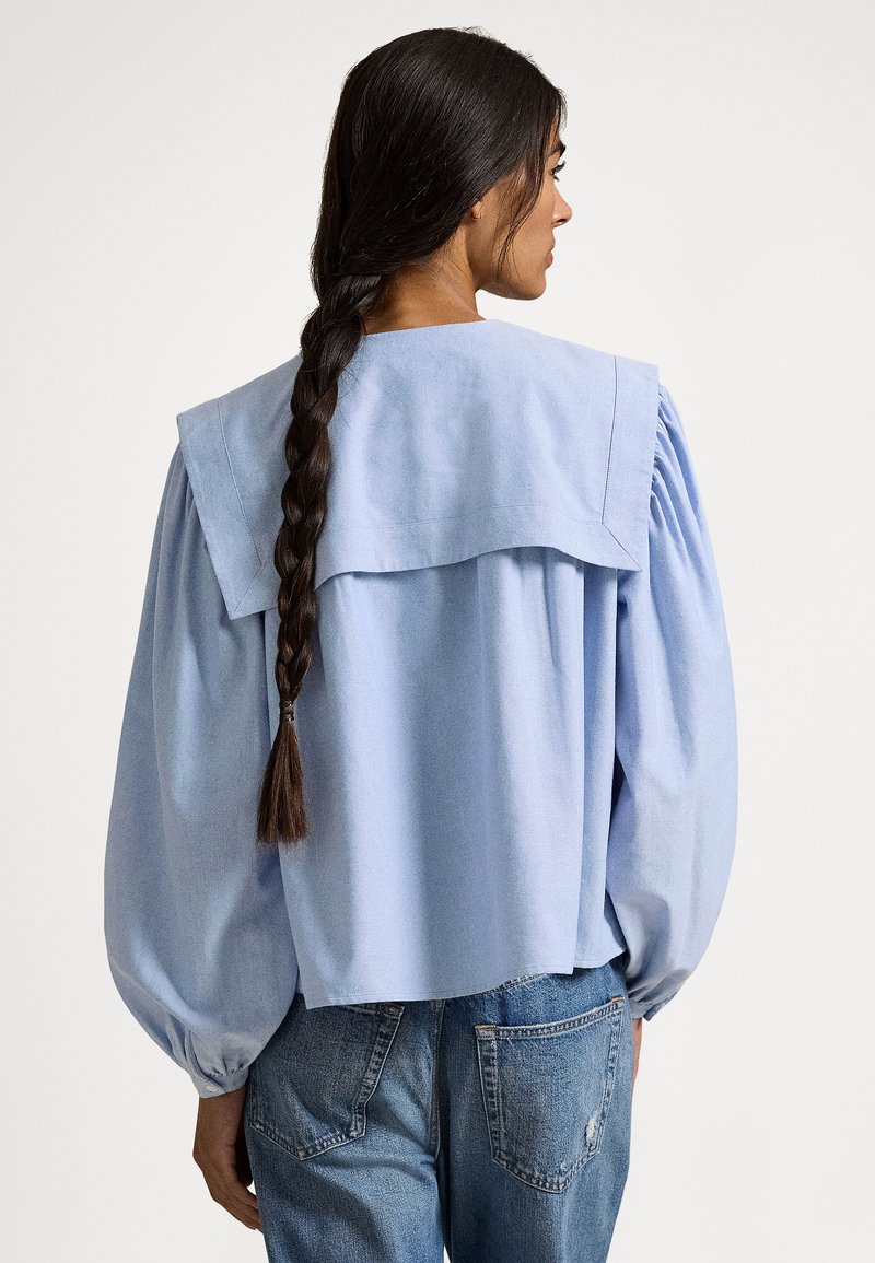 Vrouw met lang, gevlochten donker haar draagt een lichtblauw blouse met een grote matrozenkraag en pofmouwen, gecombineerd met blauwe spijkerbroek.