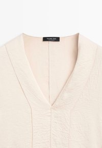 Cremfarbener V-Ausschnitt-Bluse aus strukturiertem, zerknittertem Stoff. Verfügt über eine subtile Nahtdetail und ein schwarzes Markenlabel am Hals.