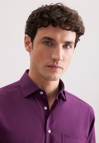 Junger Mann mit lockigem, dunklem Haar und blauen Augen, der ein lila Hemd mit Knopfleiste vor einem schlichten, hellen Hintergrund trägt.