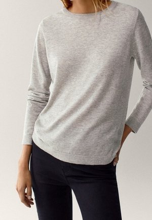 Person trägt einen hellgrauen langärmligen Pullover und schwarze Hose, steht mit einer Hand in der Tasche und der anderen entspannt an der Seite.
