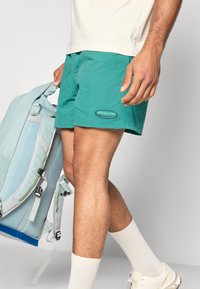 Τιρκουάζ σορτς από ελαφρύ ύφασμα, με λογότυπο σε ταμπέλα, συνδυασμένα με έναν ανοιχτό γαλάζιο σάκο πλάτης και λευκές κάλτσες μέχρι τον αστράγαλο με αθλητικά παπούτσια.