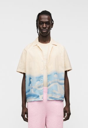 Camisa de manga corta con rayas verticales amarillas y blancas, que se desvanecen en un patrón de olas azules en la parte inferior. Lleva puesta sobre una camisa beige claro.