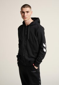 Svart zip-up hoodie i ett mjukt tyg, med en framficka och vita chevronaccenter på ärmen. Matchas med svarta joggers.