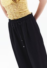Pantalones negros de corte holgado, hechos de una tela ligera, con una cintura elástica y cordón ajustable, y detalles metálicos plateados.