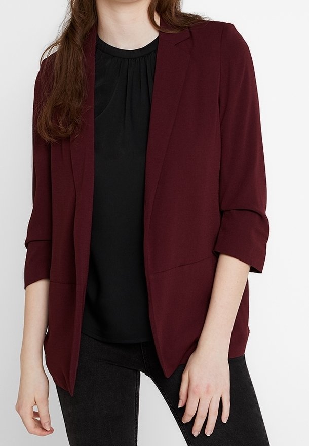 Femme portant un blazer bordeaux à manches trois-quarts sur un haut noir et un pantalon sombre, debout devant un fond blanc.