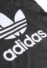 Schwarzer Stoff mit einem großen weißen Adidas-Logo, das drei Streifen und ein Trefoil-Design zeigt. Glatte Textur, minimalistisches Design.