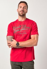 Camiseta de algodón roja con mangas cortas, que presenta un diseño impreso en gris. El modelo también lleva un reloj de pulsera marrón y pantalones verde oliva.