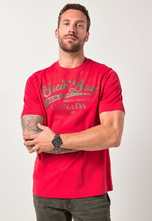 Rød bomulds t-shirt med korte ærmer, der har et trykt design i grå. Modellen bærer også et brunt armbåndsur og olivengrønne bukser.