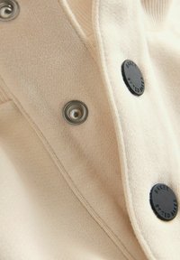 Tissu en tricot beige clair avec une texture lisse, présentant des boutons-pression noirs et des accents en métal le long du col et de la patte.