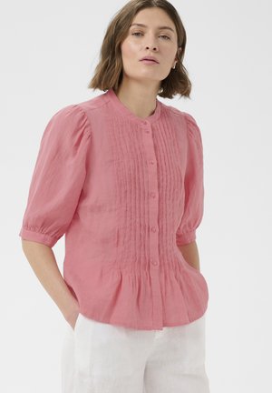 Kobieta z krótkimi brązowymi włosami, ubrana w różową bluzkę na guziki z drobnymi zakładkami oraz białe spodnie, ręce w kieszeniach, jasne tło.