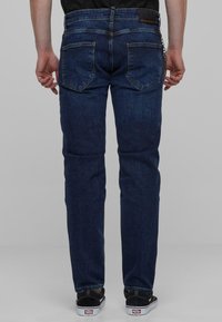 Vaqueros de mezclilla azul oscuro con un corte recto, cinco bolsillos y costuras visibles. Combinados con zapatillas negras con suelas blancas.