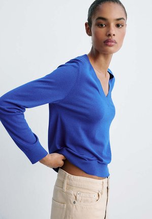 Femme aux cheveux foncés portant un pull bleu à col en V et un pantalon beige taille haute, debout avec une main sur la hanche devant un fond uni.