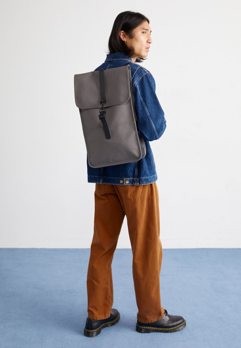 Un sac à dos gris au design structuré, avec fermeture à rabat et quincaillerie noire, porté sur une veste en denim. Un pantalon beige et des chaussures noires sont visibles.