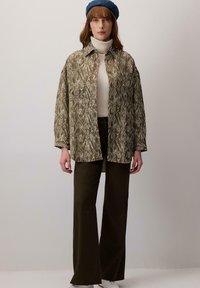 Giacca camicia verde oliva con motivo astratto bianco, abbinata a un dolcevita crema, pantaloni verde scuro e un berretto blu.