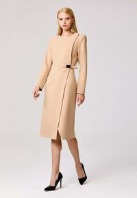 Robe beige à mi-cuisses avec manches longues, conception d'épaule structurée, fente latérale et accent de boucle noire. Associée avec des escarpins noirs.