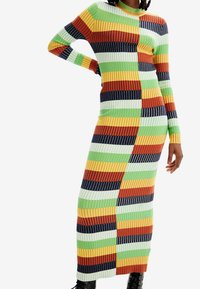 Femme portant une longue robe en maille côtelée avec des rayures horizontales vertes, jaunes, oranges, bleu marine et blanches, associée à des bottes noires.