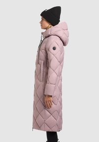 Giacca puffer rosa chiaro con motivo a rombi trapuntati, cappuccio, tasche laterali e dettagli in bottone neri. Abbinata a un berretto di maglia nero.