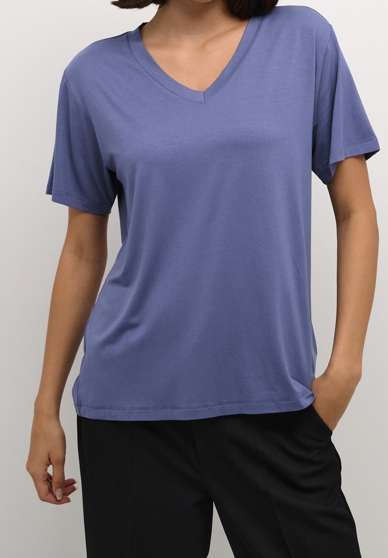 Femme portant un t-shirt violet à manches courtes, col en V, ample, et un pantalon noir, une main dans la poche, sur un fond uni.