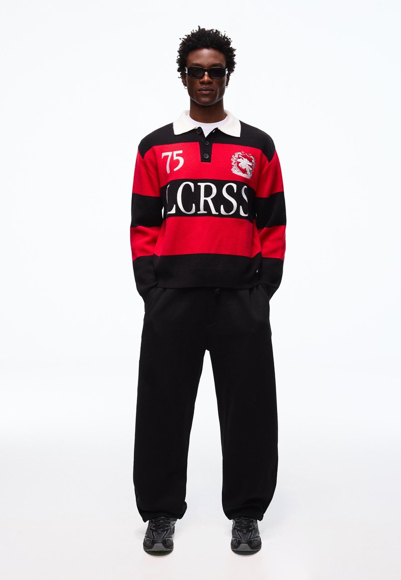 Mand iført sorte solbriller, rød og sort stribet rugbytrøje med hvid krave og nummer 75, sorte løse bukser og sorte sneakers.