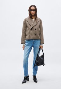 Chaqueta de lana marrón de doble botonadura, vaqueros azul claro, botas negras de tacón alto y un pequeño bolso negro. La modelo tiene el cabello largo y marrón.