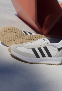 Białe i beżowe sneakersy Adidas z czarnymi paskami, widoczną podeszwą Cloudfoam, spoczywające na jasnej powierzchni z czerwonym geometrycznym tłem.