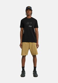 T-shirt nera G-Star con design grafico testuale, abbinata a pantaloni cargo khaki. L'individuo indossa calze nere e sneakers nere con texture.